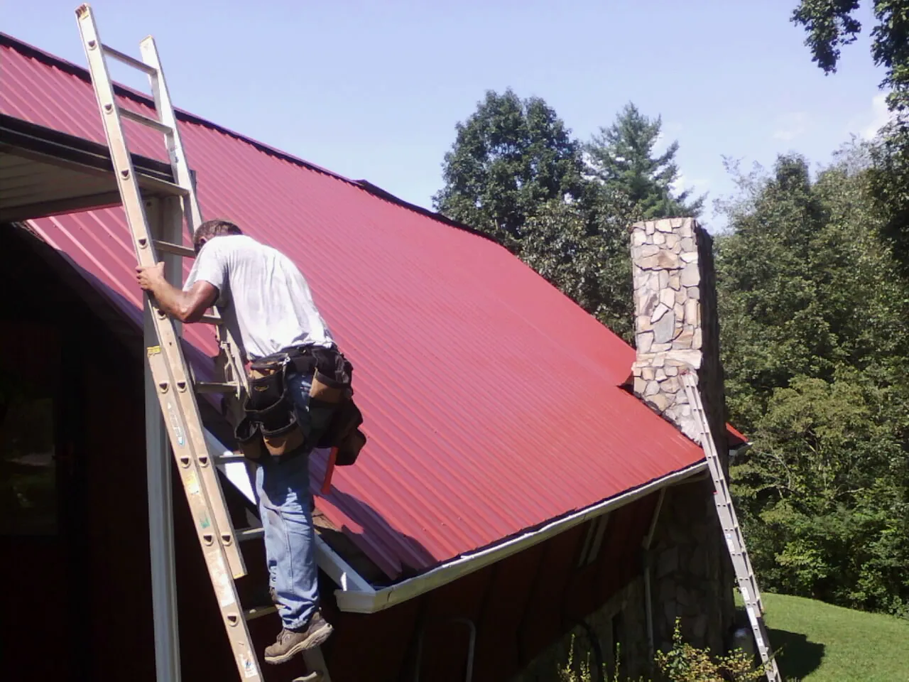 Roofer on a ladder beside a red metal roof and stone chimney — Soffit & Fascia Repair by Roofing Brothers in Tolono